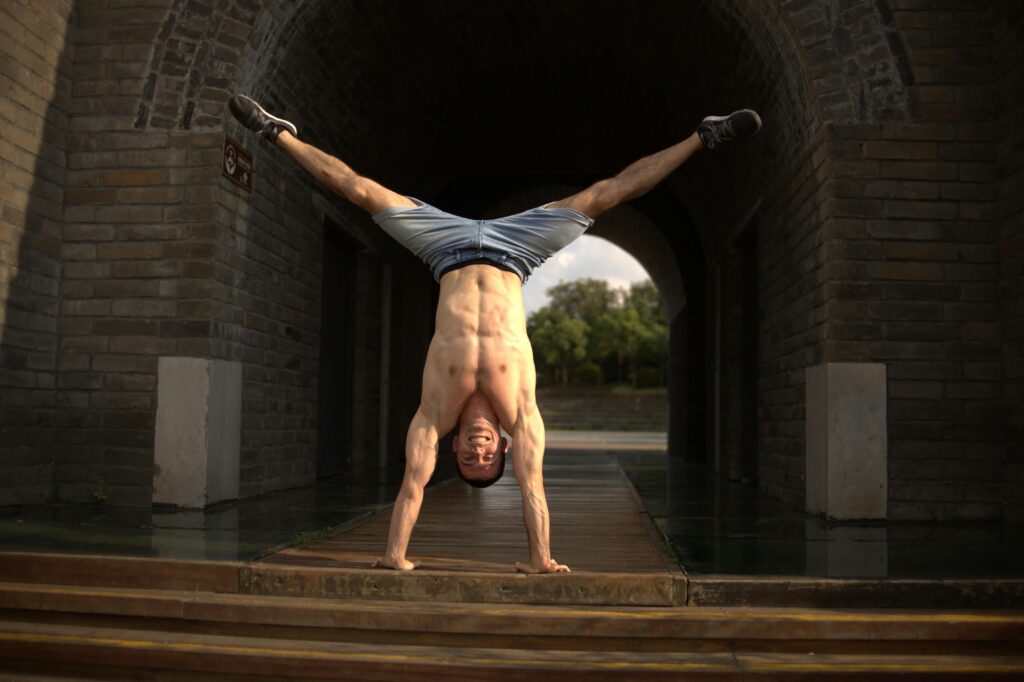 Shirtless man doing a handstand under a rustic archway, showcasing strength and balance.